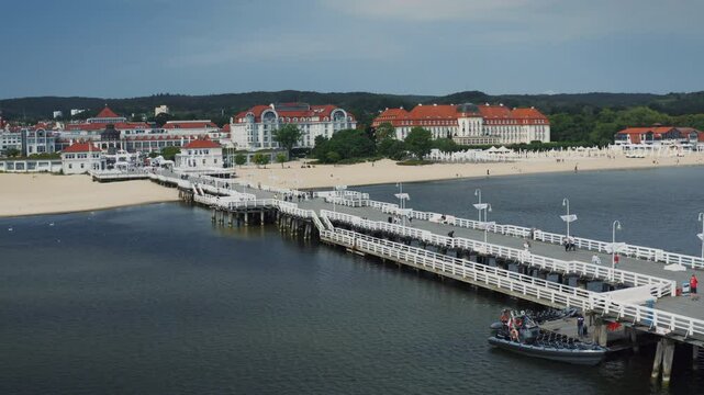 Sopot - beautiful coastal town. Long white iconic pier, sandy beach, elegant buildings featuring roofs with red tiles. People strolling on the pier. Clear sky, summer, drone shot