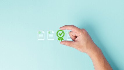 A hand holding green certificate of excellence with check mark icon surround by checklist documents with green check mark for correct and approval concept in pastel blue background.