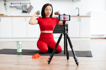 Fototapeta premium Woman in red sportswear doing home workout with dumbbells recording fitness video on smartphone. Exercise routine in kitchen interior with yoga mat, water bottle, and resistance band
