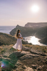 woman stands on a rocky hill overlooking a body of water. She is wearing a white dress and she is enjoying the view.