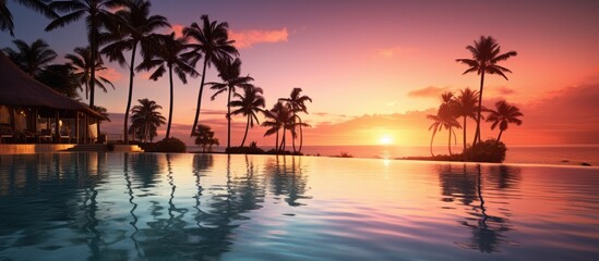Stunning sunset by the luxurious pool, palm tree silhouettes against the windy infinity pool water surface. Ideal for summer vacation with a beachfront hotel with an amazing tropical landscape.