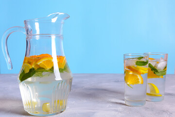 A glass pitcher filled with refreshing citrus water, ice, and mint leaves sits on a gray countertop, accompanied by two glasses of the same beverage