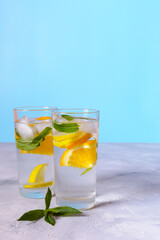 Two glasses of refreshing lemon and orange infused water are shown on a grey surface against a light blue background. Both glasses have ice cubes and fresh mint leaves