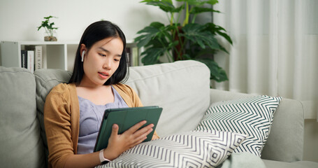 Portrait of Relaxed young Asian woman wear wireless headphones listen to music and playing digital tablet on the sofa in the living room at morning ,Free time