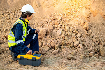 Geotechnical engineering concept. Male Asian soil engineer in reflective green vest with protective helmet and toolbox inspecting sample of laterite soil for construction improvement base road work