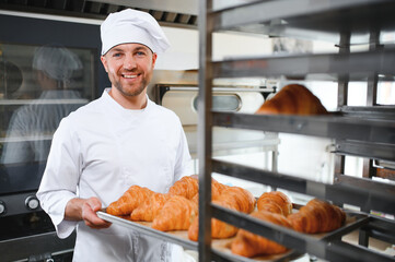 Smiling baker holding tray with fresh cooked croissants in bakehouse