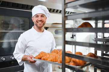 A young handsome male baker holds a tray with French croissants in front of a bakery and smiles