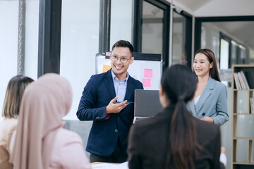 In a corporate meeting, a businessman leads a discussion on financial strategy, pointing at charts and graphs. The team collaborates on planning and analysis, using laptops and paperwork to ensure suc