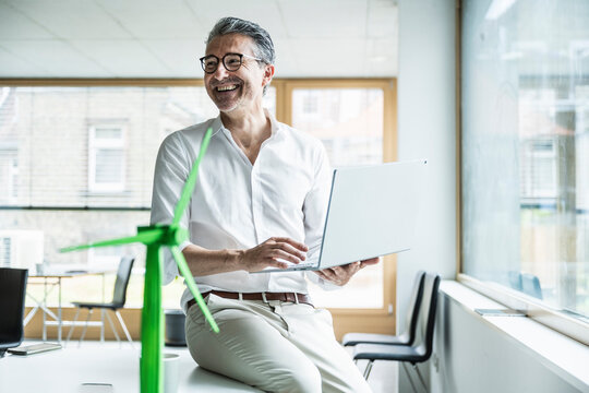 Happy businessman sitting on desk using laptop by wind turbine model in office