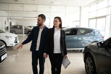 Friendly, smiling female seller showing brand new car to a customer while standing in car salon