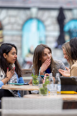 Three young women look surprised as they chat at an outdoor cafe, enjoying coffee and engaging in friendly conversation