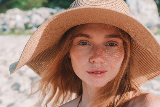 Portrait of a young attractive red-haired woman with freckles wearing a straw hat. She smiles and basks in the sun during her summer vacation, with the sun's rays falling on her face through her hat.