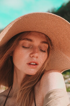 Portrait of a young attractive red-haired woman with freckles wearing a straw hat. She smiles and basks in the sun during her summer vacation, with the sun's rays falling on her face through her hat.