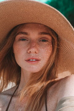 Close up of a young attractive red-haired woman with freckles wearing a straw hat. She smiles and basks in the sun during her summer vacation, with the sun's rays falling on her face through her hat.