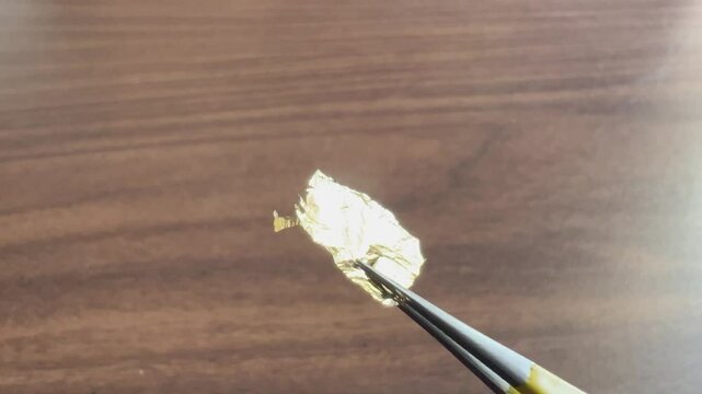 Close up fixed shot of gold leaf foil being held in fine pointed tweezers