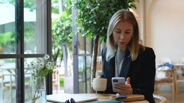 Contented businesswoman sits at cafe cozy table leisurely drinks coffee smiling looking at screen smartphone. Pleased woman takes break, enjoy beverage, reading news in phone app rest in public place.