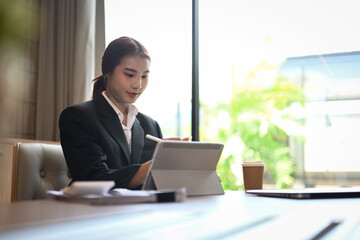 Beautiful Asian businesswoman in black suit using digital tablet at modern workplace