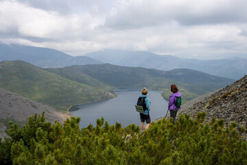 Obraz premium Two women admire a mountain lake from a mountainside. Travelers in the mountains. Traveling and hiking in the wild. Beautiful nature of Siberia and the Russian Far East. Magadan Region, Russia.