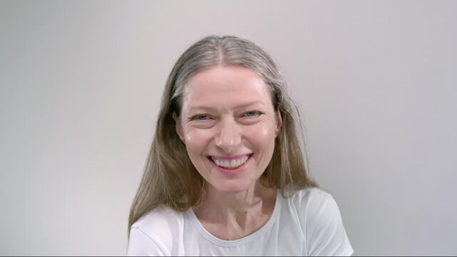 Old woman with long loose hair laughs joyfully standing alone on light background. Positive senior lady expresses real happiness emotion. Skinny healthy female pensioner has fun performing excitement