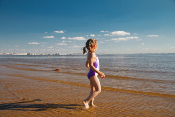 Girl of 4 years back view in swimsuit walks in sea water near city in summer