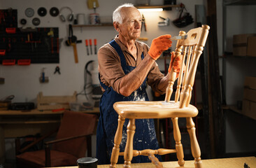 Restoration and repair of an old wooden chair. An elderly man in orange gloves, a blue apron, stands in his workshop and carefully paints furniture with a brush. © Andrii Lysenko