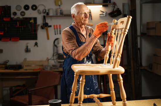 Restoration and repair of an old wooden chair. An elderly man in orange gloves, a blue apron, stands in his workshop and carefully paints furniture with a brush.