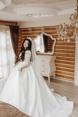 Fototapeta premium A woman in a long white dress stands in front of a mirror. She is wearing a tiara and a veil. The room is decorated with wooden furniture and a chandelier. Scene is elegant and sophisticated