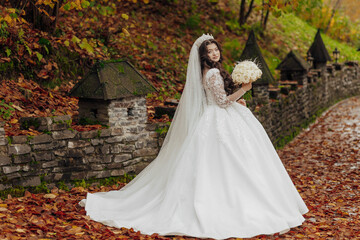 A woman in a white dress is standing in a field of autumn leaves. She is holding a bouquet of flowers and a veil