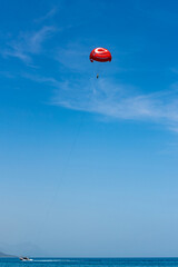A man is parasailing. The boat is towing a red parachute against a blue sky.