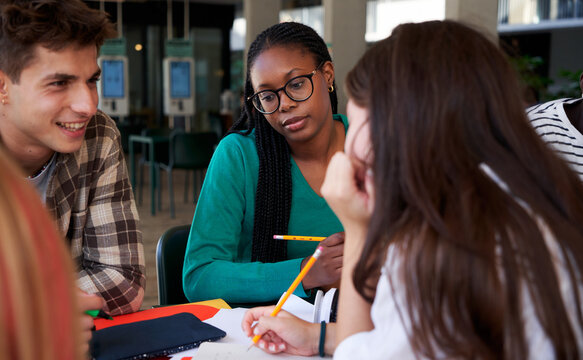Group of university students gathered in the campus bar, studying together. African girl worried about school exams surrounded by colleagues doing homework. Copy space.