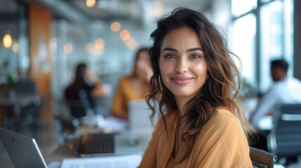 A confident woman smiles at her modern office workspace, surrounded by colleagues in a bright and collaborative environment, working on her laptop with a positive attitude.