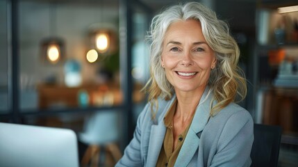 Fototapeta premium A confident senior woman with white hair and gray blazer smiling at her workspace, capturing the essence of professional success and comfort in a modern office environment.