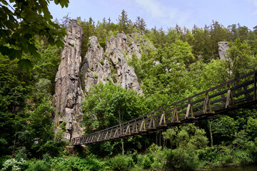 Sunny day in a lush green forest, a wooden bridge stretches across a river, leading to dramatic rock formations in Czechia