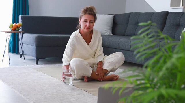 A beautiful middle-aged woman in a white outfit is drinking water afte yoga exersices