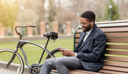 African American man is sitting on a park bench, engaged on his phone, with his bicycle nearby. The outdoor setting is calm and professional, indicating a blend of work and relaxation.