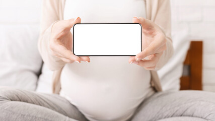 Naklejka premium A pregnant woman sits cross-legged on a bed in a bright room. She is holding a smartphone with a blank, white screen, possibly for a maternity or prenatal-related purpose, cropped