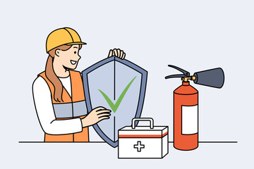 Woman safety specialist, stands near table with first aid kit and fire extinguisher and holds shield in hands. Girl safety inspector calls for caution, working to preserve life and health.