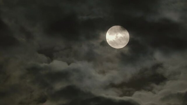 Full Moon in a dramatic night sky with dark clouds. Natural gloomy night skyscape in moonlight. Real video recording through a telephoto lens