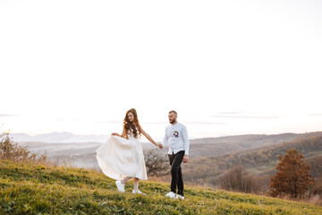 Fototapeta premium A couple is walking on a grassy hillside, hand in hand. The man is wearing a white shirt and the woman is wearing a white dress. The scene is peaceful and romantic