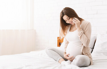 A pregnant woman is sitting on a bed, holding a cell phone to her ear while engaged in a phone conversation. She appears relaxed and comfortable as she talks on the phone in a cozy bedroom setting.