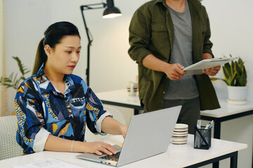 Young businesswoman working online on laptop with her colleague examining documents