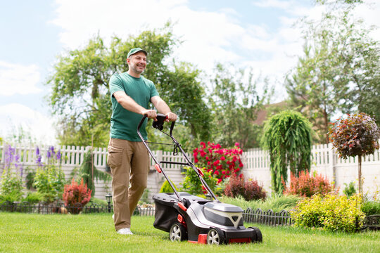 Positive man mowing the lawn using an electric lawn mower on backyard near country house, full length, free space