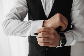 A man in a black vest and white shirt is getting dressed. He is wearing a watch and is adjusting his cuff