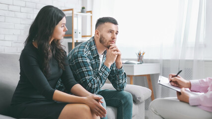 A young couple sits on a couch in a modern office setting, listening intently as a counselor takes notes. The couple appears to be discussing a personal issue, perhaps a relationship problem