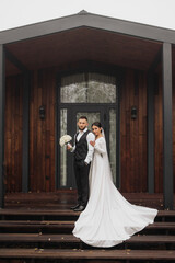 A bride and groom stand on a wooden staircase in front of a house. The bride is wearing a white dress and the groom is wearing a black suit. They are holding a bouquet and a vase, respectively