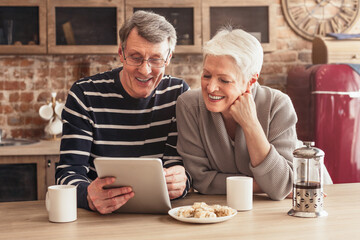 A happy senior couple are sitting in their kitchen, looking at a tablet computer. The woman has a hand on her chin and is laughing, while the man is holding the tablet and smiling