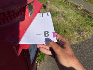 A hand puts an envelope for the United Kingdom General Election postal vote in a red post box.First Class.Envelope B