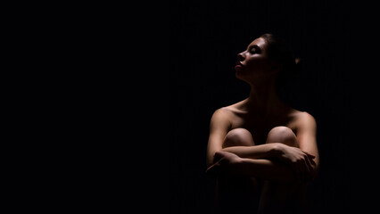 A woman sits alone in the dark, her body illuminated by a single spotlight. Her arms are wrapped around her knees, and her head is tilted up as if lost in thought, copy space