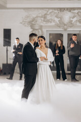 Naklejka premium A bride and groom are dancing in a foggy room. The bride is wearing a white dress and the groom is wearing a suit. There are several other people in the room, some of whom are standing
