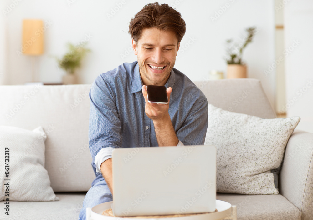 Poster A man is sitting on a couch, smiling while looking at a smartphone in his hand. He appears to be using a voice assistant to complete a task while working on a laptop.